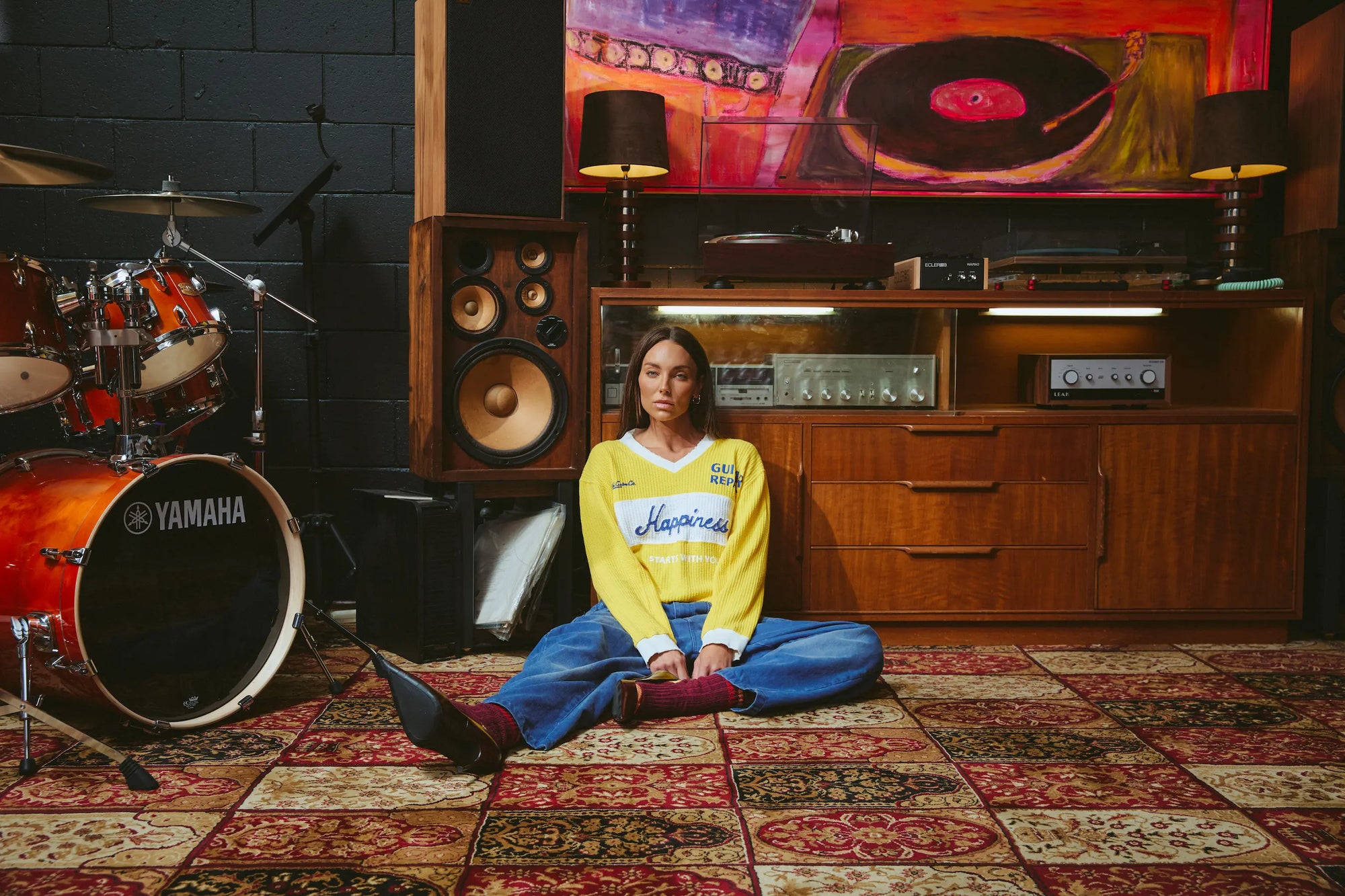 A woman sits cross-legged on a patterned rug in a music room, wearing a yellow and blue V-neck sweatshirt with 'Hershey's' written across the chest. She is surrounded by musical equipment, including a Yamaha drum set, speakers, and a wooden console with a large vinyl record painting on the wall.