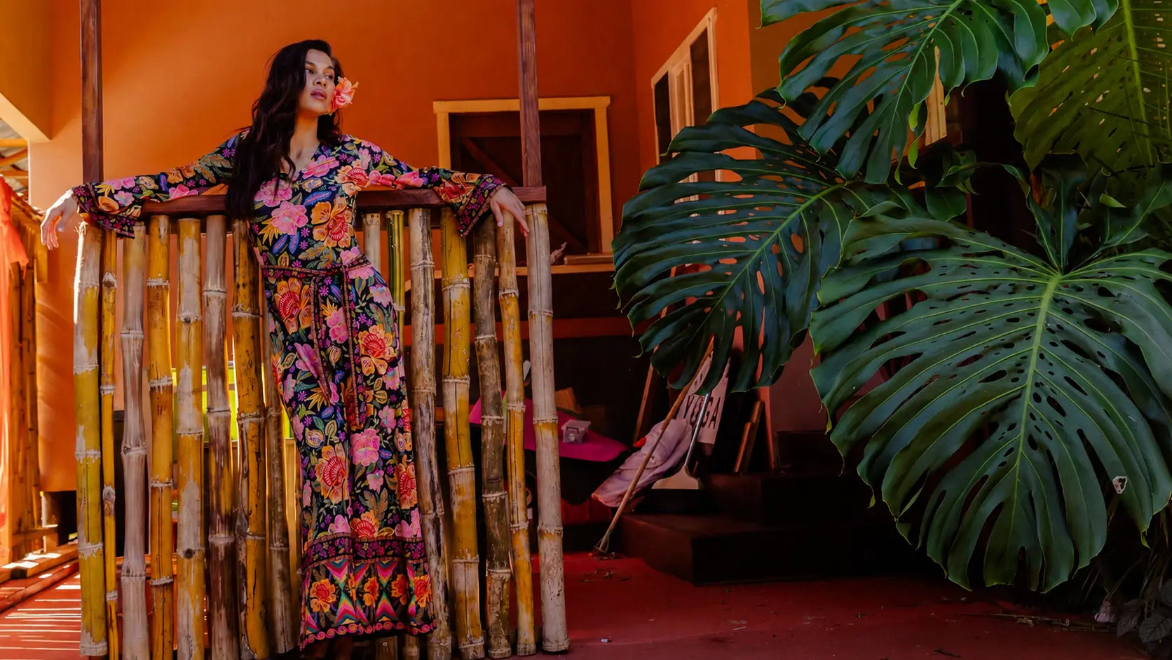 A woman wearing a colorful floral long-sleeve dress stands behind a bamboo fence in a warm-toned indoor setting with large green monstera leaves in the foreground.