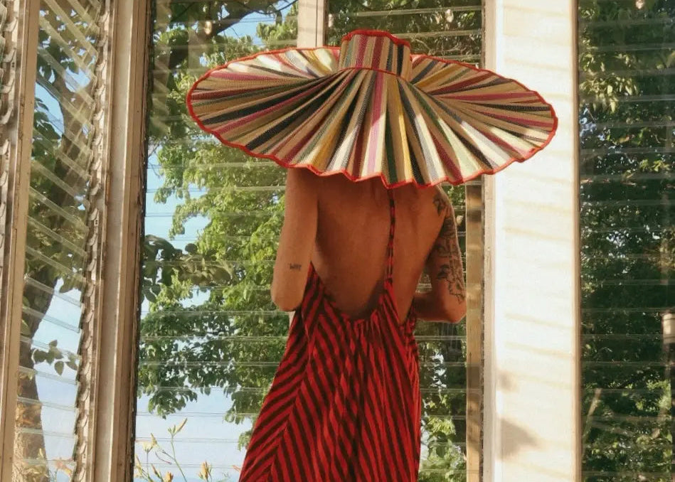 A woman stands with her back to the camera wearing a long red and black striped maxi dress and holding a large striped folding sun hat in front of her face. She is positioned on a stone patio in front of large windows with a garden view.