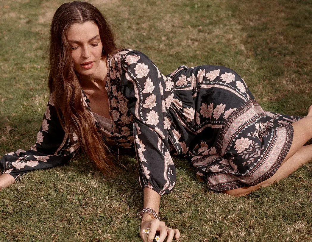 A woman lying on grass wearing a loose black maxi dress with a beige floral and geometric pattern. She is wearing a beaded bracelet and rings.
