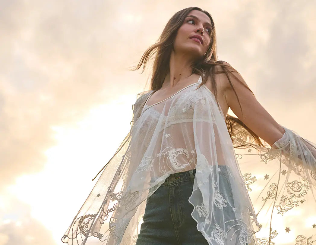A woman wearing a sheer white embroidered lace cover-up over a white camisole and dark jeans, photographed outdoors against a bright, hazy sky.