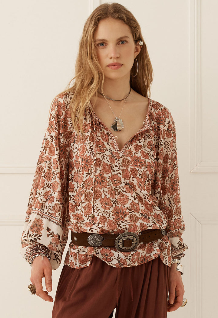 A woman modeling a long-sleeve bohemian-style blouse with a rust-brown and cream floral pattern, paired with a dark brown belt and brown pants, set against a white paneled wall.