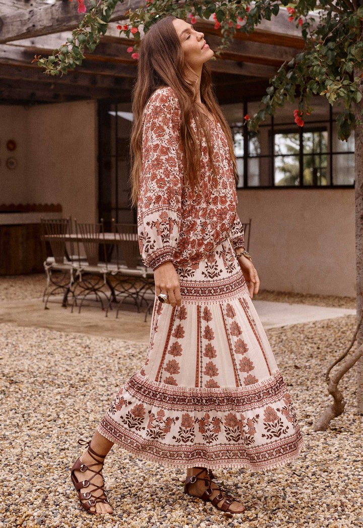 A woman wearing a bohemian-style two-piece dress set with a rust brown and cream floral pattern, walking outdoors on a gravel path near a patio.