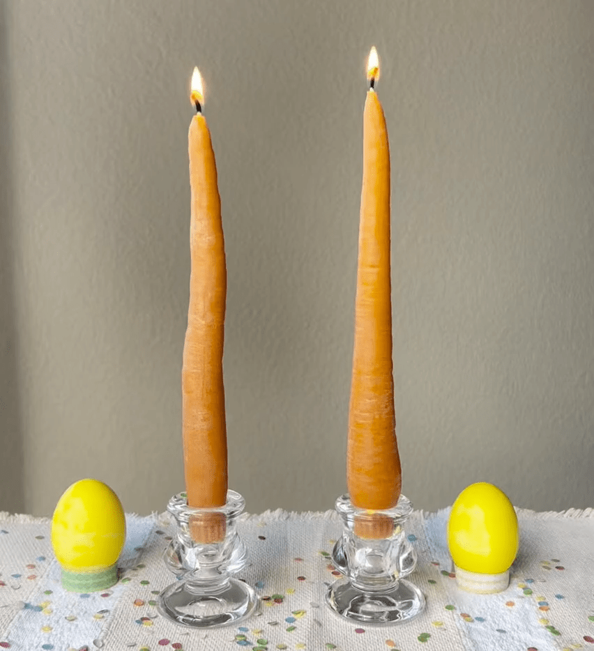 Two lit orange taper candles in glass holders on a tablecloth with yellow eggs.