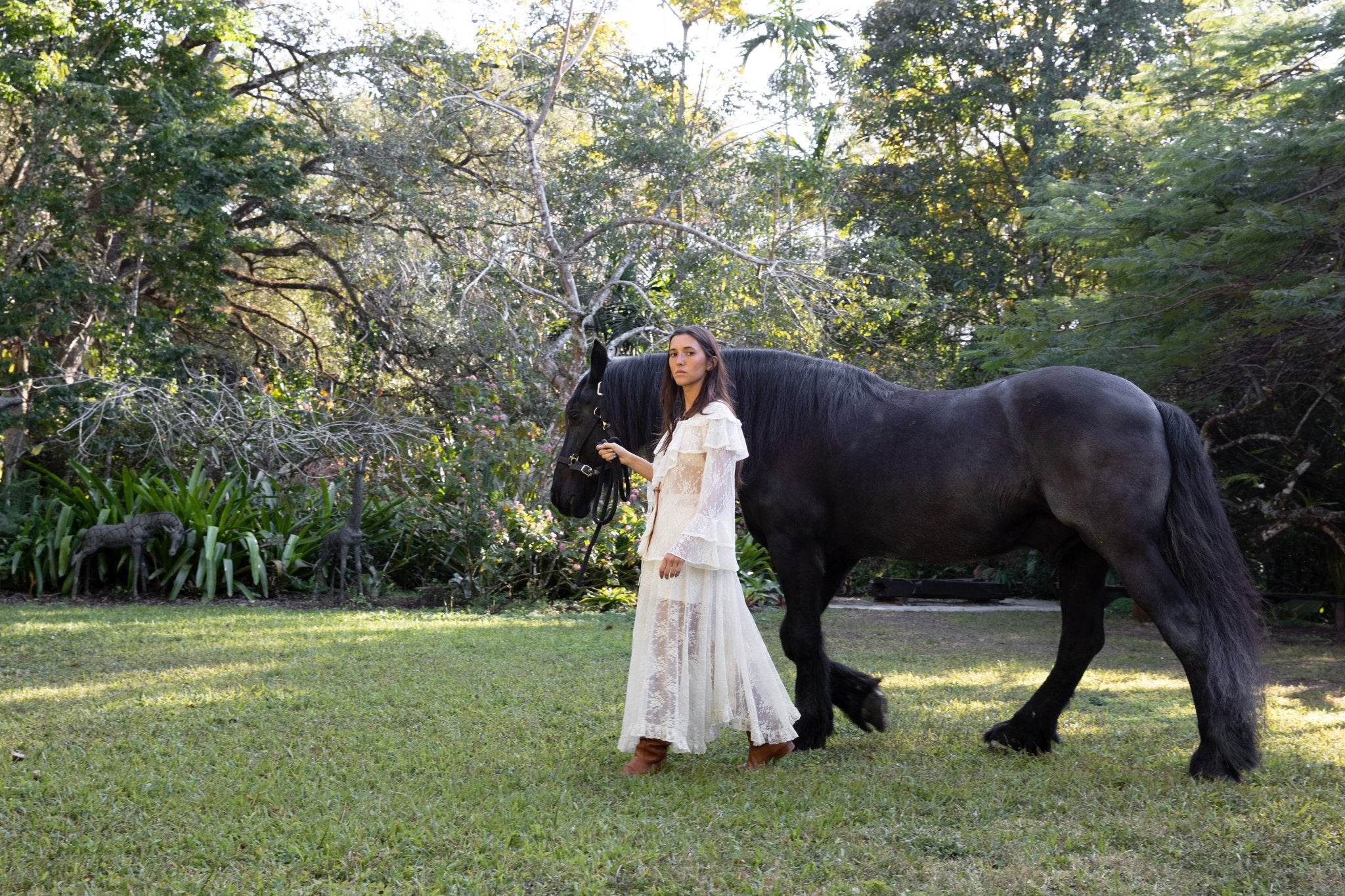 Woman in a white dress standing next to a black horse in a grassy area with trees.