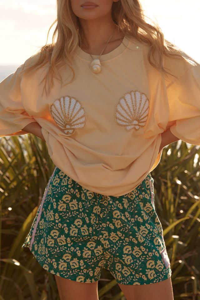 Woman wearing a light-colored top with shell designs and green floral shorts in a natural setting.