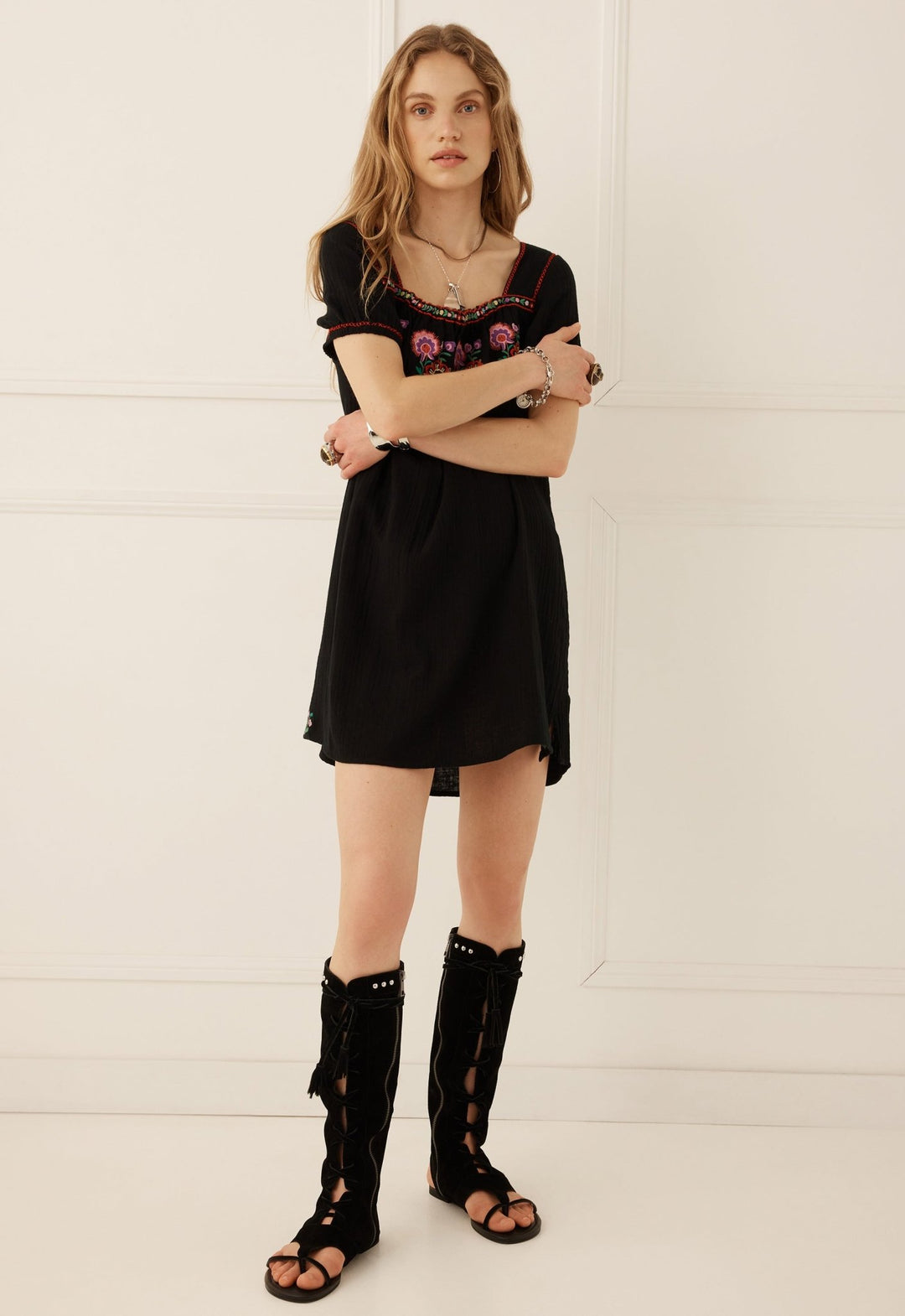 A woman modeling a black short-sleeve dress with colorful floral embroidery on the bodice, paired with black lace-up gladiator sandals, standing against a white paneled wall.