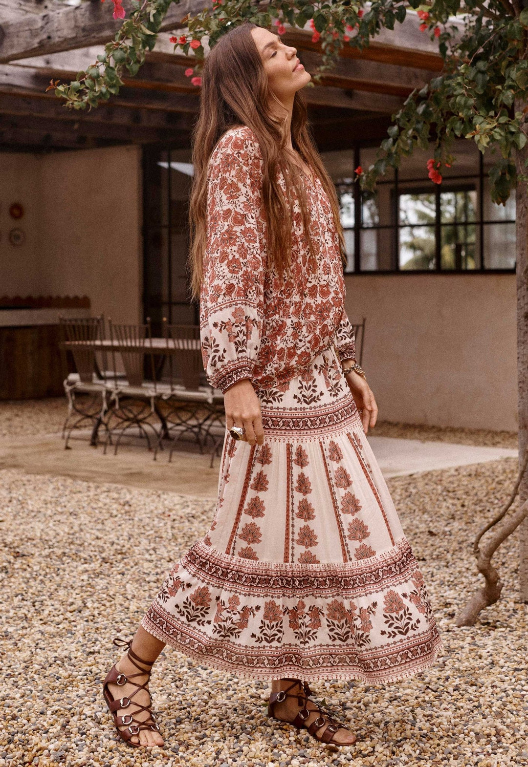 A woman wearing a bohemian-style two-piece dress set with a rust brown and cream floral pattern, walking outdoors on a gravel path near a patio.