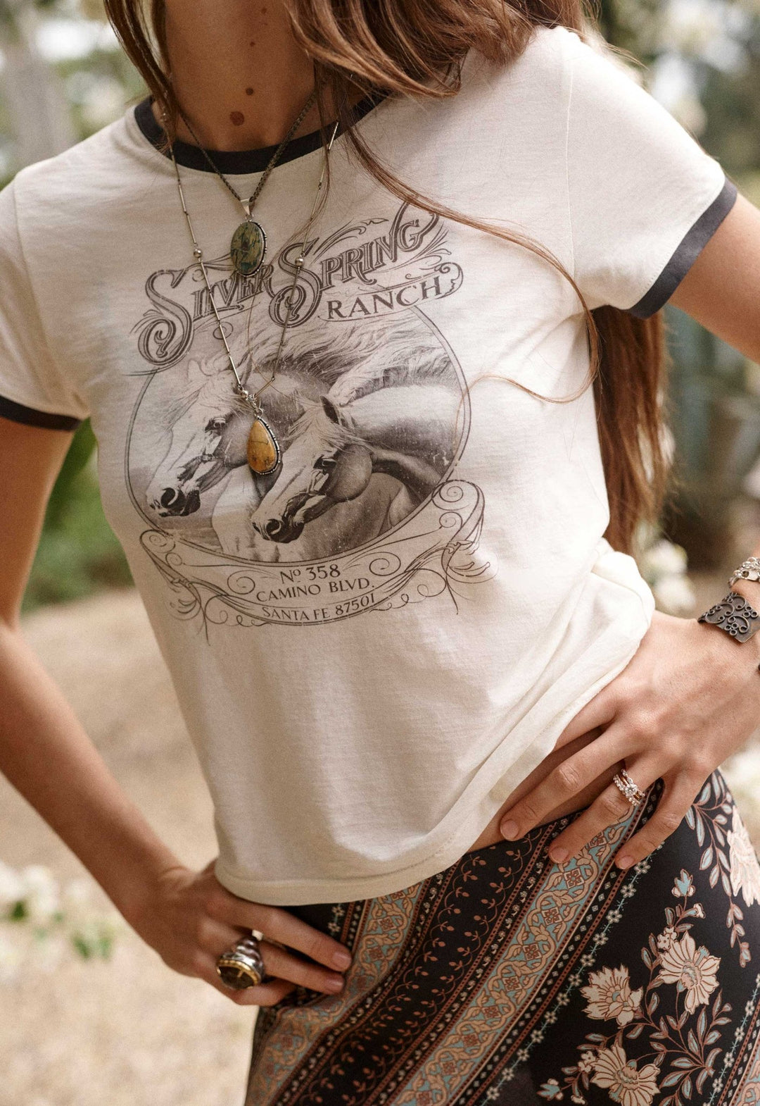 A woman wearing a cream-colored ringer t-shirt with black trim and a vintage 'Silk Spring Ranch' graphic print on the front, paired with a dark patterned skirt.